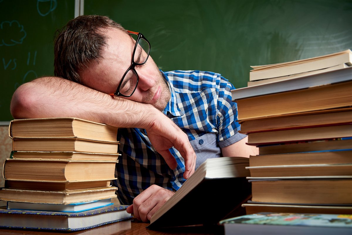Homem adormecido em cima de uma pilha de livros, exausto de tanto estudar sobre o exame do sono